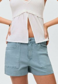 Svijetloplave pamučne kratke hlače s prednjim džepovima i kopčanjem na dugme, u kombinaciji s bijelim, teksturiranim majicom bez rukava. Detalji uključuju v-izrez.