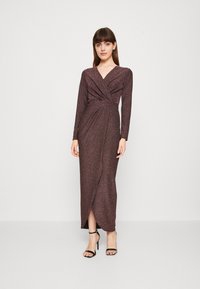Robe métallique longue et ajustée en violet foncé avec un design à l'avant torsadé, des manches longues et une fente latérale. Assortie avec des escarpins noirs à bride de cheville.