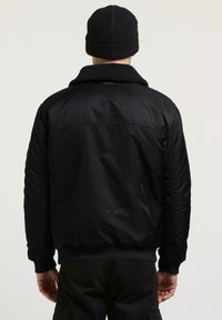 Giacca bomber nera con colletto foderato in pile e texture. Presenta polsini e orlo a coste, e un tessuto liscio con una leggera lucentezza.