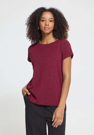 Femme aux cheveux bouclés porte un haut bordeaux à manches courtes et un pantalon noir, debout sur un fond clair uni, une main dans la poche.