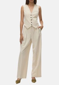 Gilet beige à rayures et pantalons à jambes larges. Le gilet a un col en V, six boutons et une poche. Tissu lisse avec un léger brillant.