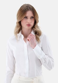Weißes Langarmshirt mit klassischem Kragen, ausgestattet mit Knopfmanschetten und einer glatten Textur. Das Shirt wird mit einem hellen Taillenbund getragen.