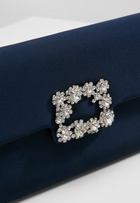 Námořnická modrá saténová kabelka s obdélníkovou přezkou zdobenou krystaly na předním klopě.