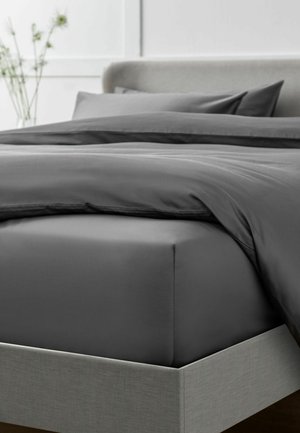 Bett mit grauem Spannbettlaken, dunkelgrauem Bettbezug und Kissen, in einem minimalistischen Schlafzimmer mit einer verschwommenen Blumenvase im Hintergrund.