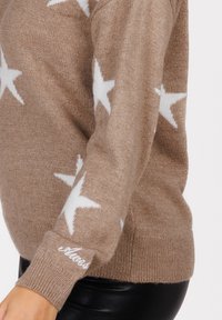 Beige gestrickter Pullover mit weißen Sternmustern an Ärmeln und Körper, mit Logo-Stickerei am Bündchen, getragen mit schwarzen Lederhosen.
