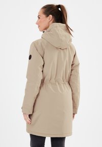 Beige takki, jossa on kiristysvyötärö, huppu ja elastiset hihansuut. Vasemmassa hihassa on logopatch. Suora, vettä hylkivä kangas.