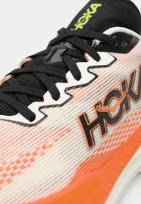 Primeiro plano de um sapato atlético com atacadores pretos, parte superior de malha branca e laranja, e logotipo "HOKA" em preto e laranja na lateral.
