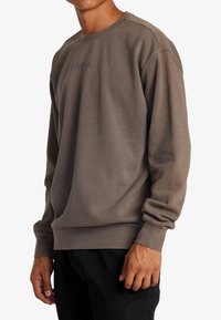 Brun crewneck-tröja gjord av mjukt tyg. Har ribbade muddar och fåll samt en subtil logotryck på bröstet.