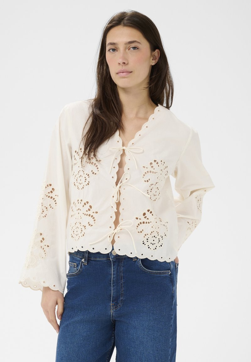 Femme portant un chemisier crème avec une broderie ajourée florale et des fermetures à nouer, associé à un jean taille haute bleu foncé.