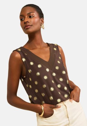 Femme portant un top marron sans manches à col en V avec motif de rayons de soleil dorés, bracelet en or, boucles d'oreilles en forme de soleil et pantalon clair.