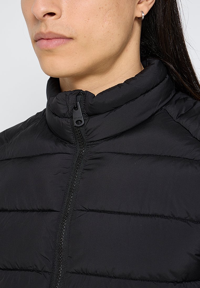 Chaqueta de plumas negra con cuello alto, que presenta un cierre frontal y un diseño acolchado para aislamiento. Textura de tela suave, hardware mínimo.