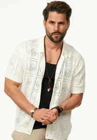Weißes, kurzärmliges Strickshirt mit offenen Mustern, mit Knopfleiste vorne und lockerer Passform, über einem schwarzen Tanktop getragen.