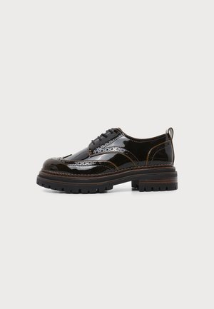 Zapato brogue de cuero negro brillante con suela gruesa y dentada, costuras detalladas, perforaciones y cierre de cordones, mostrado de lado sobre fondo blanco.