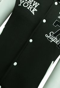 Zwarte jas met witte drukknoopjes, geborduurde tekst "NEW YORK" en "Super" detail. Zachte stof met een gladde textuur, minimalistisch ontwerp.