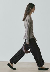 Szara bluzka z długim rękawem, czarne luźne spodnie i czarne baleriny. Trzyma małą brązową torebkę w kształcie półksiężyca. Neutralne tło.