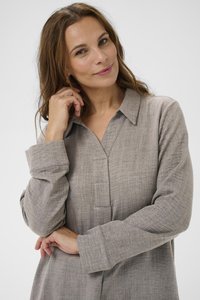 Kvinna med långt brunt hår som bär en lös beige texturerad blus, som försiktigt rör vid sitt ansikte och ler mjukt mot en neutral bakgrund.