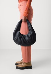Personne portant un pantalon corail et des bottes beige tenant un grand sac à main noir en forme de croissant par sa poignée sur le côté.