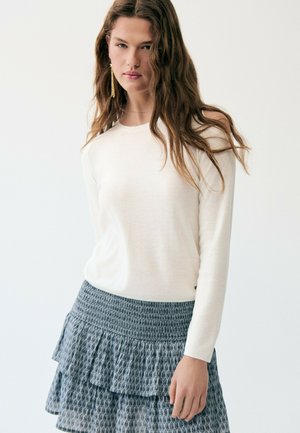 Junge Frau mit langem braunem Haar, die ein weißes langärmliges Oberteil und einen blau gemusterten Stufenrock trägt, vor einem schlichten hellen Hintergrund stehend.