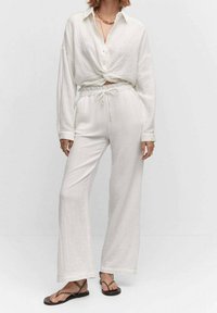 Witte linnen tweedelige outfit bestaande uit een geknoopte overhemden en wijde broek met een elastische tailleband en koord. Platte sandalen.