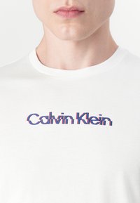Balta medvilnės marškinėliai su apvaliu kaklu, puikuojantys daugiatkoniu "Calvin Klein" logotipu raudona ir mėlyna spalvomis, šiek tiek iškreipta dizaino forma.