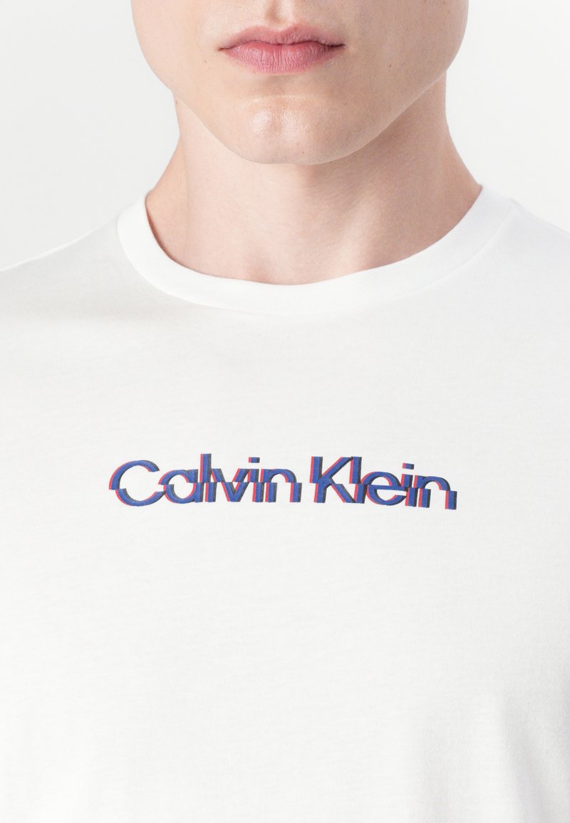 Camiseta de algodón blanca con cuello redondo, que presenta un logotipo de "Calvin Klein" en colores rojo y azul, con un diseño ligeramente distorsionado.