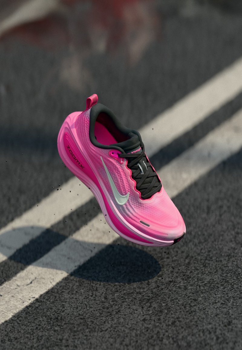 Φωτεινό ροζ και μαύρο παπούτσι τρεξίματος Nike με ασημένιο λογότυπο σε ενδιάμεση πτήση πάνω από άσφαλτο δρόμο με λευκή διαχωριστική γραμμή.