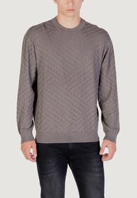 Pull en tricot gris avec un motif géométrique texturé, col rond et poignets et ourlet côtelés. Porté sur un jean foncé.