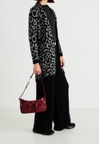 Cardigan de estampado de leopardo negro y gris, top negro acanalado, pantalones de pierna ancha negros, sosteniendo un bolso burdeos con dos bolsillos frontales.