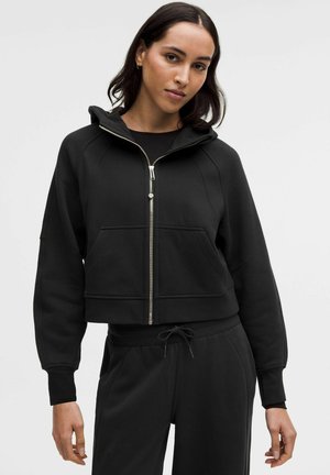 Zwarte cropped hoodie gemaakt van zachte stof met een ritssluiting aan de voorkant, grote zakken en een trekkoord onderaan. Voorzien van raglanmouwen en een capuchon.