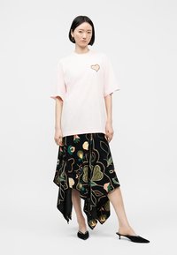 Γυναίκα που φοράει μια oversized μπλούζα σε απαλό ροζ χρώμα με μικρό σχέδιο καρδιάς, συνδυασμένη με ασύμμετρη μαύρη φούστα με πολύχρωμο λουλουδένιο σχέδιο.