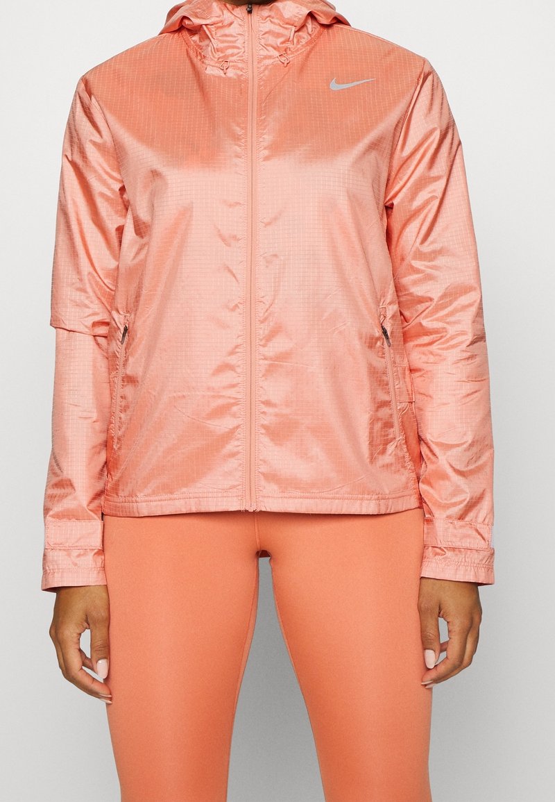 Veste coupe-vent Nike corail clair avec fermeture éclair avant et poches zippées, portée avec des leggings corail assortis.