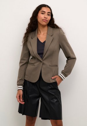 Braun taillierter Blazer mit gestreiften Manschetten, zwei Fronttaschen, kombiniert mit schwarzen, gefalteten Leder-Shorts, vor einem neutralen Hintergrund.