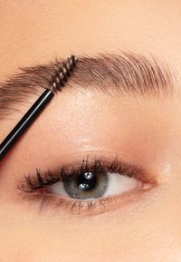 Czarny pędzelek do brwi ustawiony na tle brązowych i ciemnych włosów, podkreślający pojedyncze włoski brwi; widoczny jest delikatny odcień skóry i niebiesko-zielone oko.