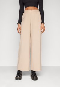 Pantalons larges en tissu beige clair avec une texture lisse et une fermeture à bouton. Associés à des bottes noires à lacets.