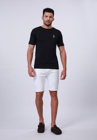 T-shirt noir à manches courtes avec un petit motif blanc sur la poitrine gauche, associé à un short en denim blanc et des chaussures à enfiler marron.