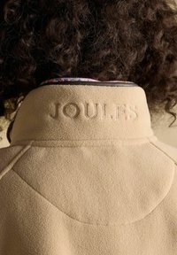 Asmuo su garbanotais plaukais dėvi smėlio spalvos vilnonį švarkelį su ant nugaros apykaklės išsiuvinėtu užrašu „JOULES“, matyti švarkelio tekstūra ir siuvimo detalės.