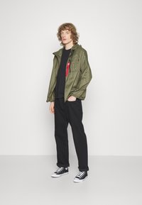 Olive green lightweight jacket with a hood, zippered front, and chest pocket; paired with a black shirt and loose black pants, featuring black sneakers.