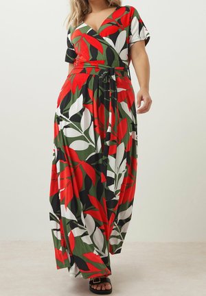 Kobieta w długiej, wielokolorowej sukience kopertowej z czerwonym, zielonym, czarnym i białym motywem liści, w połączeniu z czarnymi sandałami z odkrytymi palcami.