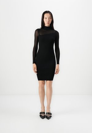 Black ribbed dress with a high neck and long sleeves; fitted silhouette; smooth texture; paired with pointed-toe black heels featuring strap accents.