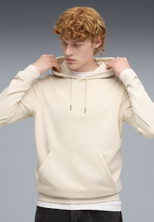 Young man adjusting cream-colored hoodie with front pocket and white printed Puma Athletics logo on chest against gray background.