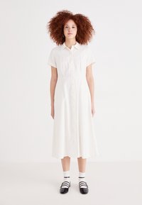 Rochie albă cu mâneci scurte și fustă evazată, realizată dintr-un material ușor. Asociată cu pantofi negri și șosete albe. Design simplu.
