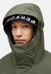 Olive green hooded jacket with a high collar, featuring a black inner hood displaying white text. Smooth, water-resistant fabric.