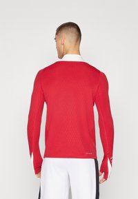 Piros, hosszú ujjú sportpóló bordázott textúrával, fehér gallérral és hüvelykujjnak való nyílásokkal az ujjaknál. A hátsó nézet az illeszkedő dizájnt és a márkázást mutatja.