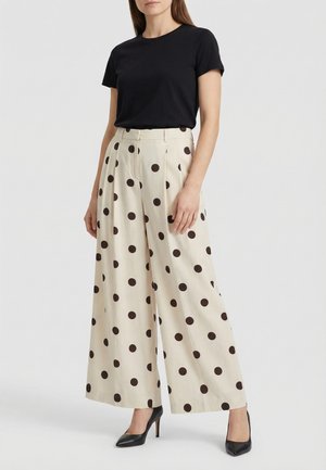 Donna che indossa un top nero a maniche corte, pantaloni larghi color crema a vita alta con grandi pois neri e scarpe nere con tacchi alti, in piedi.