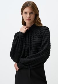 Zwart getextureerde lange mouw top met een hoge hals, met een geplooid ontwerp en licht gepofte mouwen. Draagt met zwarte hoge taillebroek.
