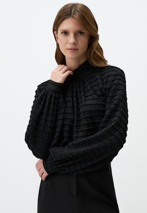 Maglia nera a maniche lunghe con collo alto, caratterizzata da un design plissettato e maniche leggermente a palloncino. Indossata con pantaloni neri a vita alta.