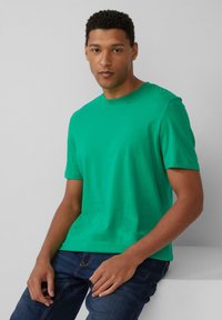 Zielona bawełniana koszulka z okrągłym dekoltem i krótkim rękawem, z delikatnym logo na piersi. Połączona z ciemnymi dżinsami.