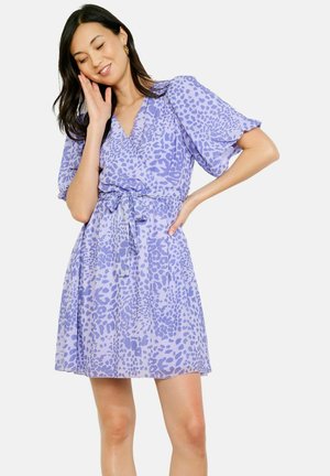 Mujer vistiendo un vestido lila con estampado de leopardo morado, de manga corta y con cinturón, posando con una mano en la cadera y la otra cerca de su rostro.