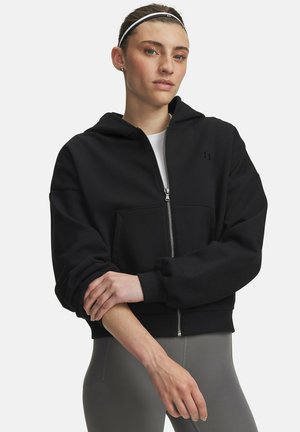Jonge vrouw met hoofdband draagt een zwart zip-up hoodie en grijze legging, met gekruiste armen kijkend naar de camera.