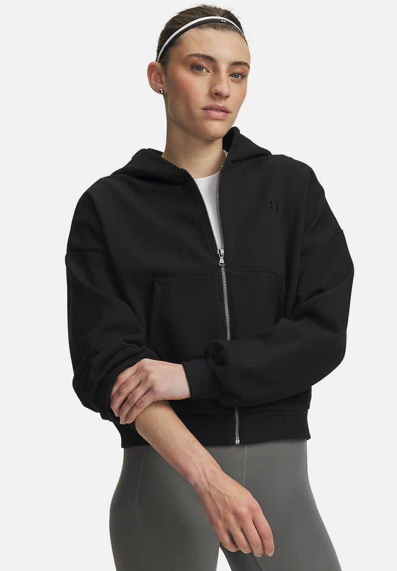 Jeune femme avec un bandeau portant un sweat à capuche noir zippé et des leggings gris, bras croisés et regardant la caméra.